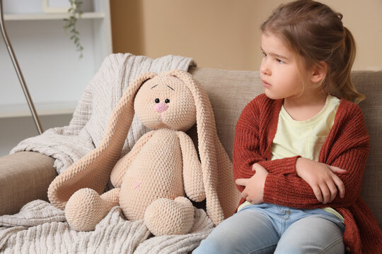 Sad little girl with toy bunny sitting on sofa at home