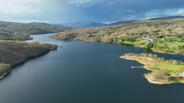 Aerial view of Encoro de Prada reservoir with forested hills and a small village under a cloudy sky in A Veiga, Ourense, Spain.