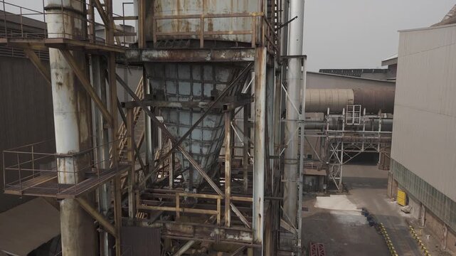 Shot in D-Log M for professional color grading, Aerial view of large industrial factory with rusty metal structures and cooling pipes under a hazy grey sky