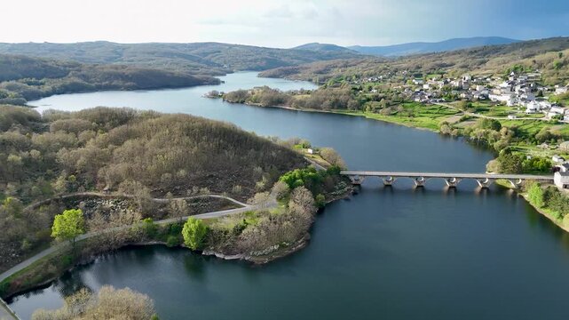 Aerial view of Encoro de Prada reservoir and its arched bridge surrounded by lush green hills near the village of A Veiga, Ourense, Spain.