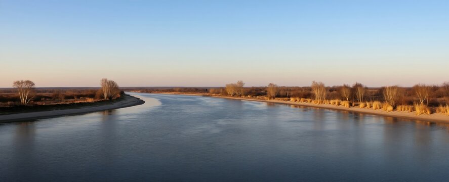 Wide river flows toward sea under clear sky. Sandy banks with sparse trees and dry grass line the water. Landscape is calm and serene during daytime.