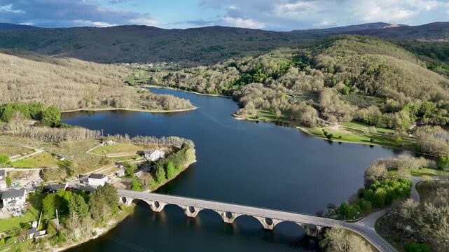 Aerial view of the stone arched bridge over Encoro de Prada reservoir surrounded by lush green hills and small houses A Veiga, Ourense, Spain.