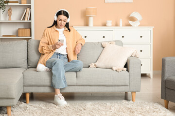 Obraz premium Young woman in headphones listening to audiobook with mobile phone on sofa at home