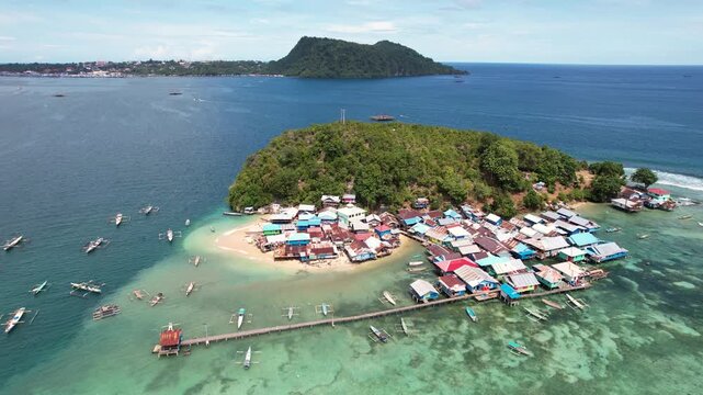 Aerial view of a traditional stilt village built over turquoise waters with outrigger boats and lush green hills in Jayapura, Papua, Indonesia.