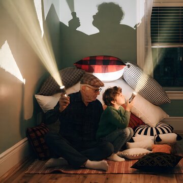 Grandparent and grandchild build a lopsided pillow fort in the living room, flashlight shadows and playful chaos. Cozy family scene for childhood joy.
