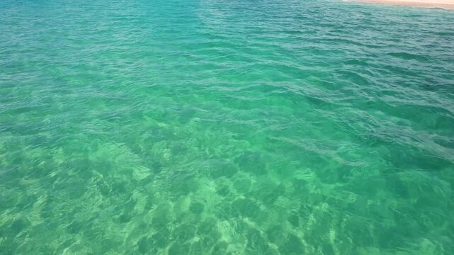 Aerial view of the crystal clear turquoise sea water with gentle ripples over a sandy bottom under bright sunlight in Paralia Ofriniou, Greece.