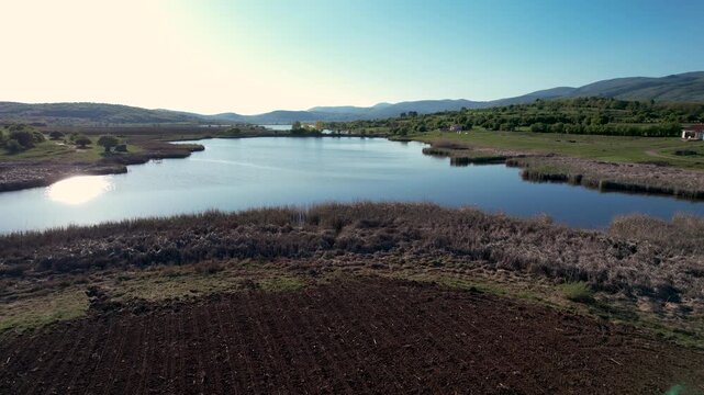 Aerial view of Smilovci Lake with its calm waters surrounded by reeds, rolling green hills, and a freshly plowed field under a clear blue sky in Smilovci, Pirot District, Serbia.