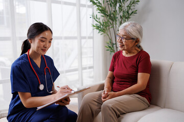 Fototapeta premium Home healthcare professional in blue scrub taking notes on a clipboard during a medical consultation with a senior asian woman sitting comfortably on a sofa, providing support and care