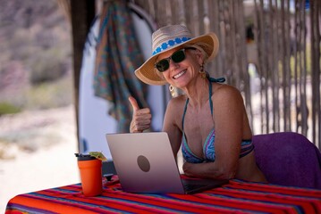 Fototapeta premium Mature woman works on a computer at a tropical beach, showing joy while enjoying her vacation in a sunny location