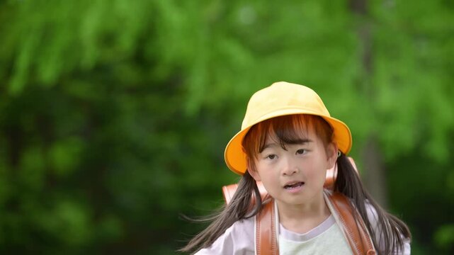 新緑の中ランドセルで走る登校する笑顔の女の子　黄色い帽子を被った1年生の新生活と通学や学校、学資保険や未来を見つめるなどのイメージのスローモーション動画