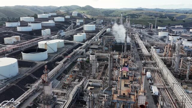 Phillips 66 San Francisco Refinery in Rodeo, California, with large American flag waving over industrial pipes and white storage tanks under overcast sky. Aerial drone shot.