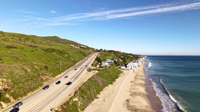 Pacific Coast Highway traffic flows past Malibu Beach as the drone tracks alongside cars driving the iconic California coastal route between the ocean and green hills. Corral Beach view.