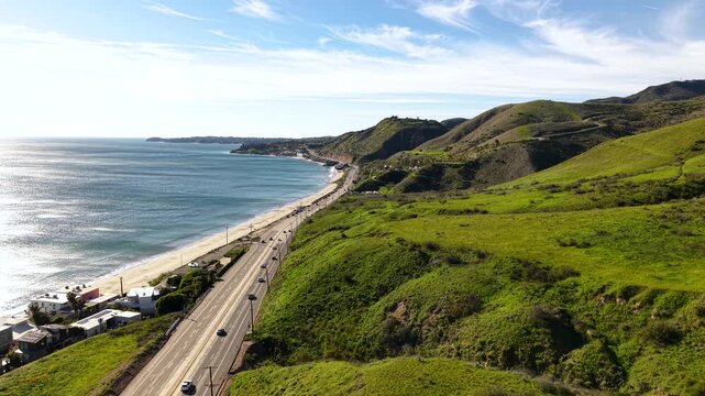 Lush green cliffs rise above Pacific Coast Highway as the drone strafes sideways along the Malibu coastline, capturing rare verdant California landscape after winter rains.