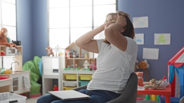 Middleaged hispanic woman teacher rubbing temples then typing on laptop in building classroom filled with toys, tent, and stuffed animals; stress burnout work balance.