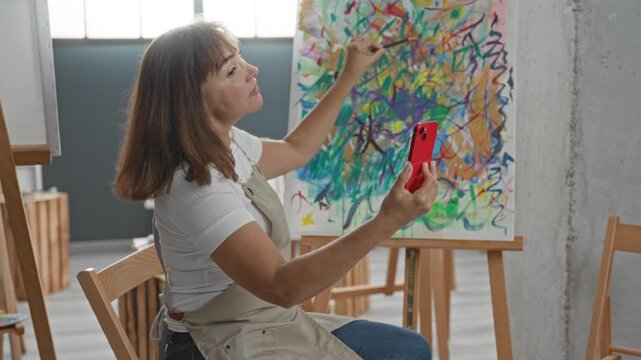 Woman artist holding red smartphone and paintbrush while painting a spontaneous abstract canvas on an easel in an art studio; quiet creativity.