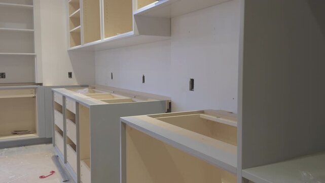 Interior view of a modern kitchen during cabinet installation. Unfinished light natural wood cabinet carcasses with gray painted frames and panels are shown in various stages of assembly.