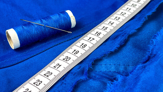 A close-up of sewing supplies on torn blue fabric. A spool of thread, a needle, and a measuring tape suggest crafts, tailoring, and textile manufacturing.