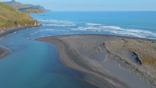 Aerial view of the coastline at Marokopa, New Zealand. The shot shows the river meeting the ocean, with waves crashing on the black sand beach. Rolling hills are in the background.