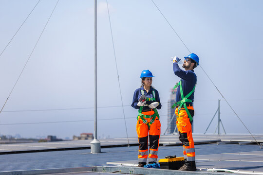 Two solar engineers in safety uniform and harnesses working on rooftop solar farm, Maintenance team of technicians inspecting solar panels installation on industrial solar roof under blue sky