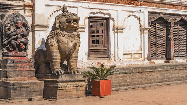 Stone sculpture of a lion sitting on a pedestal. Mouth open, big teeth. On the wall is a bas-relief of a god Hanuman, eviscerating a demon Hiranyakashipu. Barred windows. Nepal. Kathmandu. Durbar
 