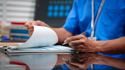Healthcare worker in blue scrubs reviewing paperwork on clipboard at hospital desk, holding pen, clinical setting with lab supplies and monitor in background. © NanSan