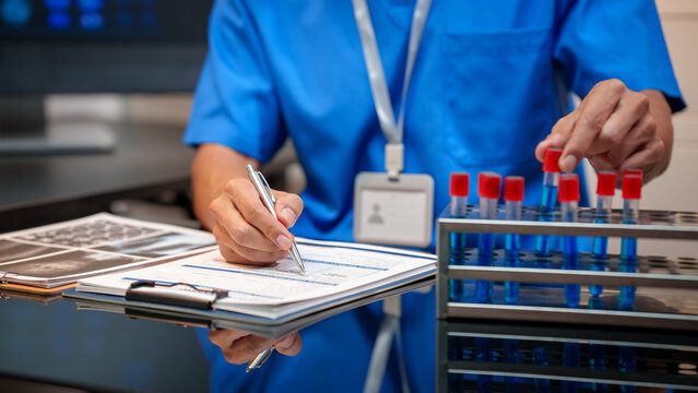 Medical lab technician in blue scrubs writing on clipboard beside rack of blood test tubes in laboratory, healthcare diagnostics concept.