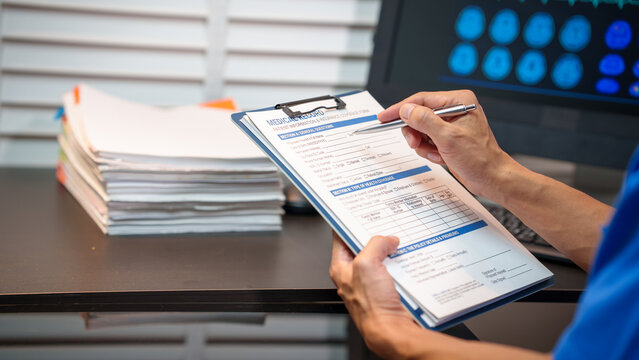 Person reviewing medical record form on clipboard at desk with stack of paperwork, pen in hand, and computer monitor showing brain scan images in a clinic or office setting.