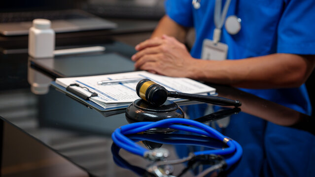 Medical legal concept: judge gavel and stethoscope on desk with nurse or doctor in background, clipboard paperwork and blurred office setting.