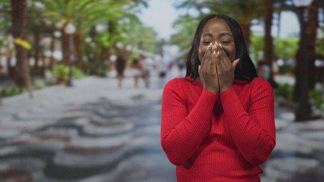 Woman covers mouth with hands, smiling in red sweater on a palm lined street with blurred pedestrians and patterned pavement; surprise reaction.