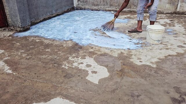 A worker applies white URP liquid coating to a concrete terrace floor for waterproofing. Using a broom and bucket, they spread the sealant along a gray wall over the damp, stained concrete surface.