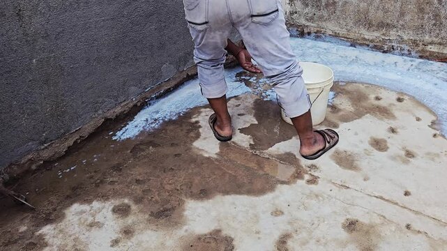 A worker applies white URP liquid coating to a concrete terrace floor for waterproofing. Using a broom and a bucket, they spread the sealant over the damp, stained concrete surface, textured gray wall