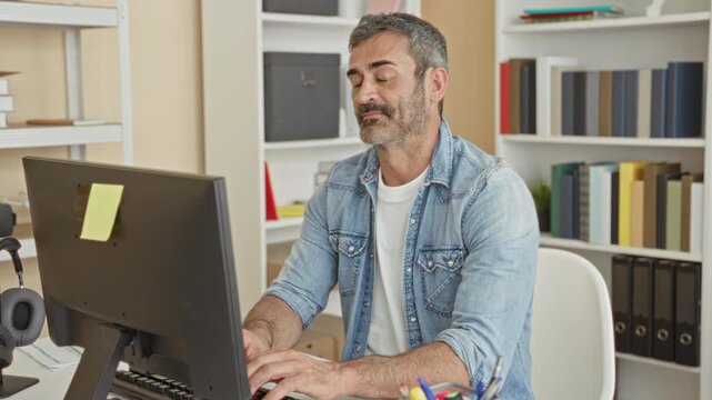 Man at computer using hands and fingers to gesture thumbs up and type at desk in building office with bookshelves and binders visible; confidence approval.