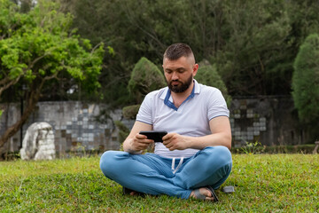 Obraz premium Man sitting cross-legged on green grass, focused on a smartphone in his hands, surrounded by a landscaped garden featuring neatly trimmed shrubs and trees in the background