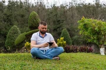 Obraz premium Man with a beard sitting cross-legged on green grass in a city park, holding a smartphone, with lush trees and shrubs providing a serene backdrop in an outdoor nature environment