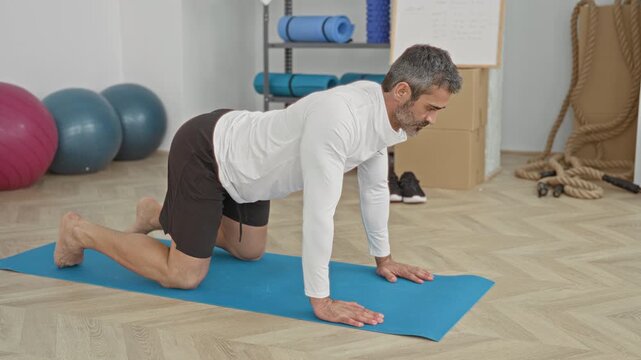 Man kneeling on a blue yoga mat in a studio, hands planted and back arching in a stretching pose with exercise balls nearby; calm focus.