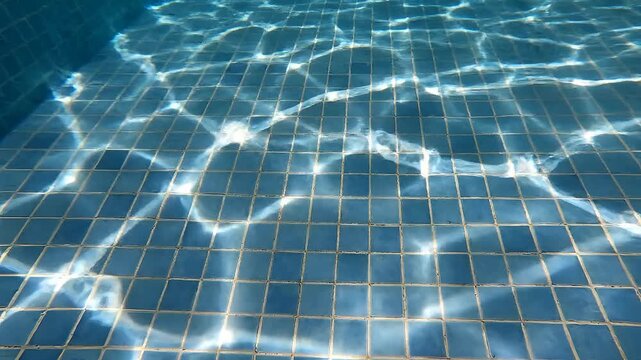 Underwater view of a swimming pool with tiled bottom and sun reflections.