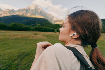 Obraz premium Woman enjoying outdoor landscape with headphones, hiking past mountains, casual backpack, serene mood, bright natural light, lifestyle moment in open field and meadow