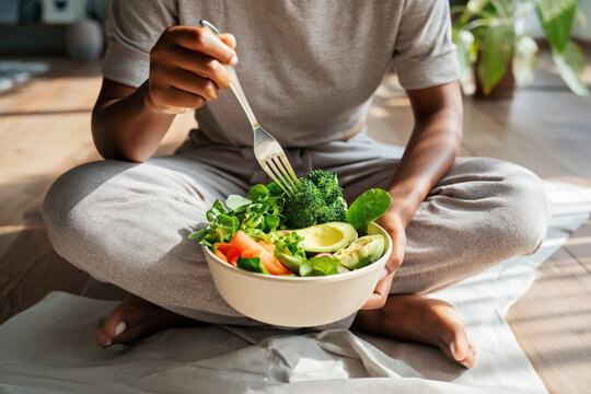 a person sitting on the floor with their legs crossed, holding onto an eco-friendly bowl filled with vibrant and colorful vegetables like broccoli, carrots, avocados, and leafy greens