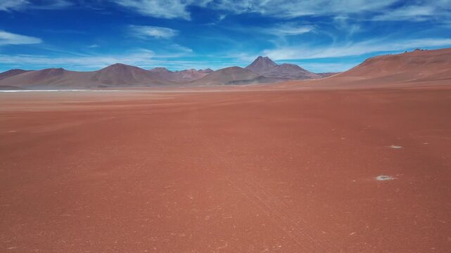 Dolly shot over vast red desert plain with salt patches gliding toward dramatic volcanic mountain range under vivid blue sky with wispy clouds. San Pedro de Atacama, Chile