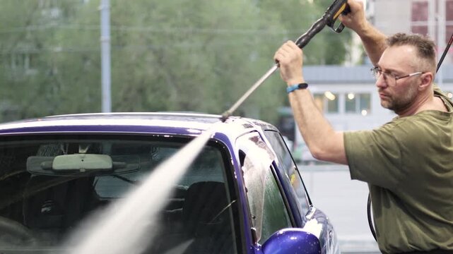 Join a man in this cheerful video as he washes his blue car at a self-service car wash. The combination of sun, soap, and water brings a burst of freshness to his day. Slow motion 4k video