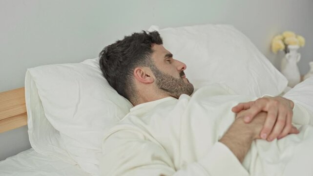 Man lying on bed with hands clasped on stomach and eyes closed, pillow and wooden headboard visible in a bedroom; restless sleep.