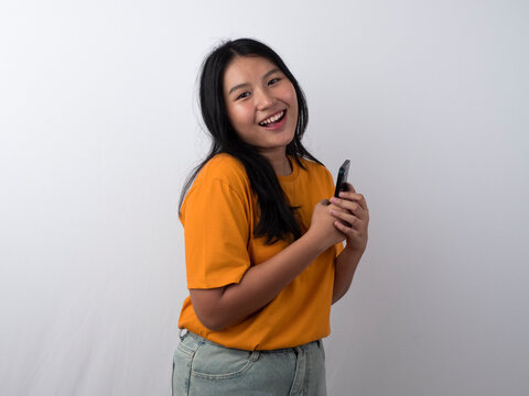 Joyful young Asian woman in yellow shirt holding a smartphone while showing a happy expression, isolated by white background.