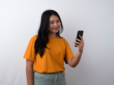 Joyful young Asian woman in yellow shirt holding a smartphone while showing a happy expression, isolated by white background.