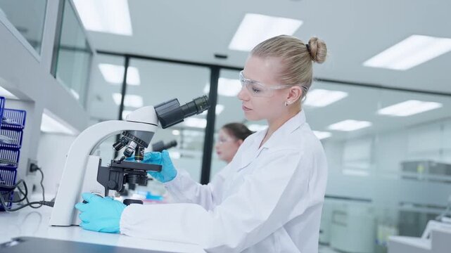 A scientist in a laboratory examines a test tube while using a microscope. Another researcher is working nearby. The focus is on the process of scientific research and discovery.