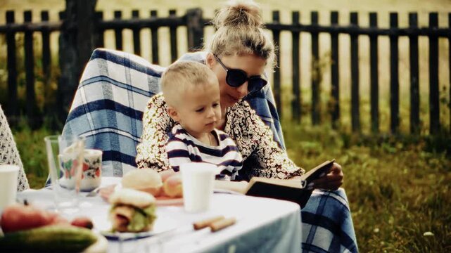 Mother Reading Book to Toddler during Outdoor Picnic. Family Lunch in Garden. Weekend Recreation and Leisure 