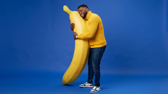 Joyful man in yellow sweater hugging a giant banana with a smile against vibrant blue backdrop, playful surreal food scene with oversized fruit and cheerful humor