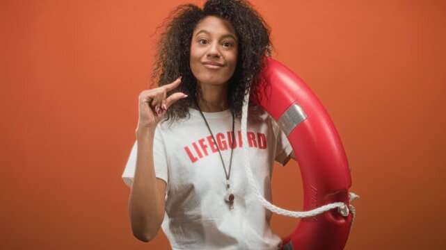 Woman lifeguard pinches fingers while holding red lifebuoy and whistle necklace in studio; safety confidence.