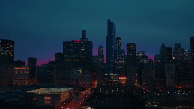 Stunning aerial panning shot of downtown Chicago cityscape at night, revealing illuminated skyscrapers and busy streets under vibrant purple and blue twilight sky.