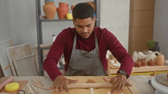 Man potter rolling clay with hands and rolling pin on worktable in studio, wearing apron and focused gaze; quiet concentration.
