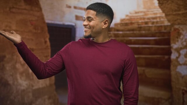 Man with beard in maroon shirt holds palm up and smiles by a stone stairway in a building; contentment.